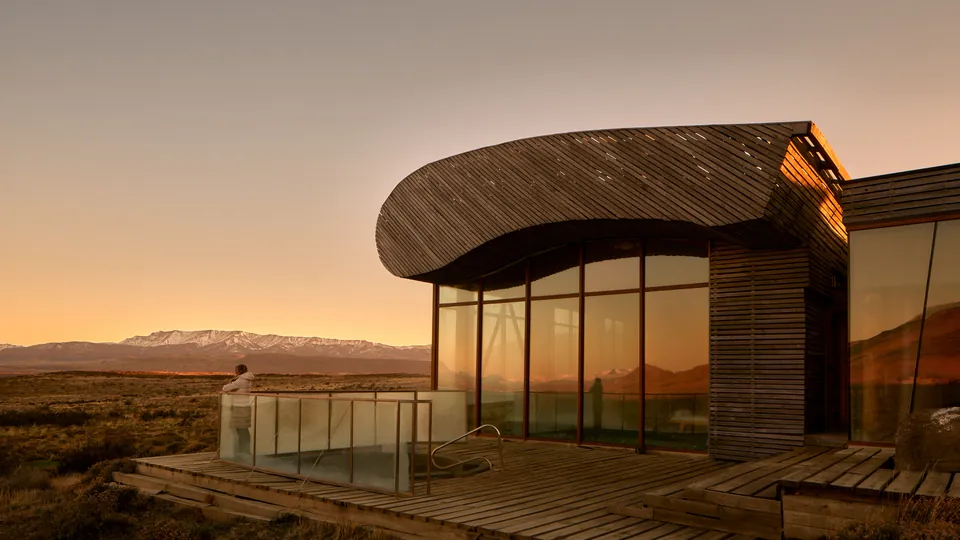 Curved timber lodge at dusk, Patagonian steppe and a snow-capped range behind