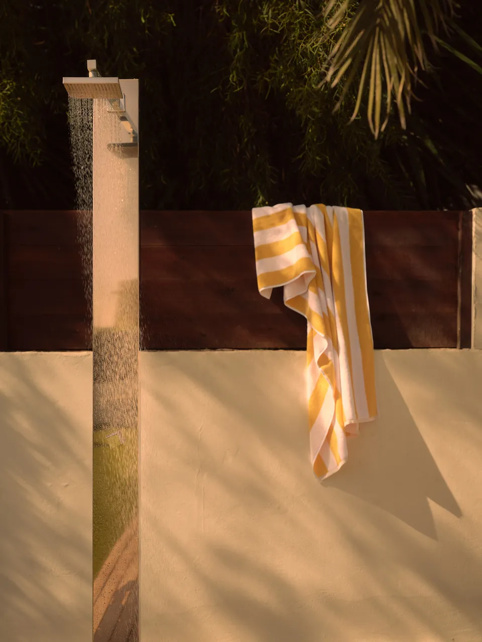 Outdoor shower against a sunlit wall, a striped towel hung to dry