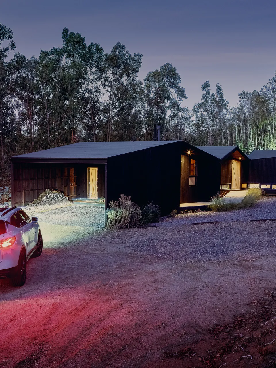 Black cabins at dusk, headlights on the gravel drive