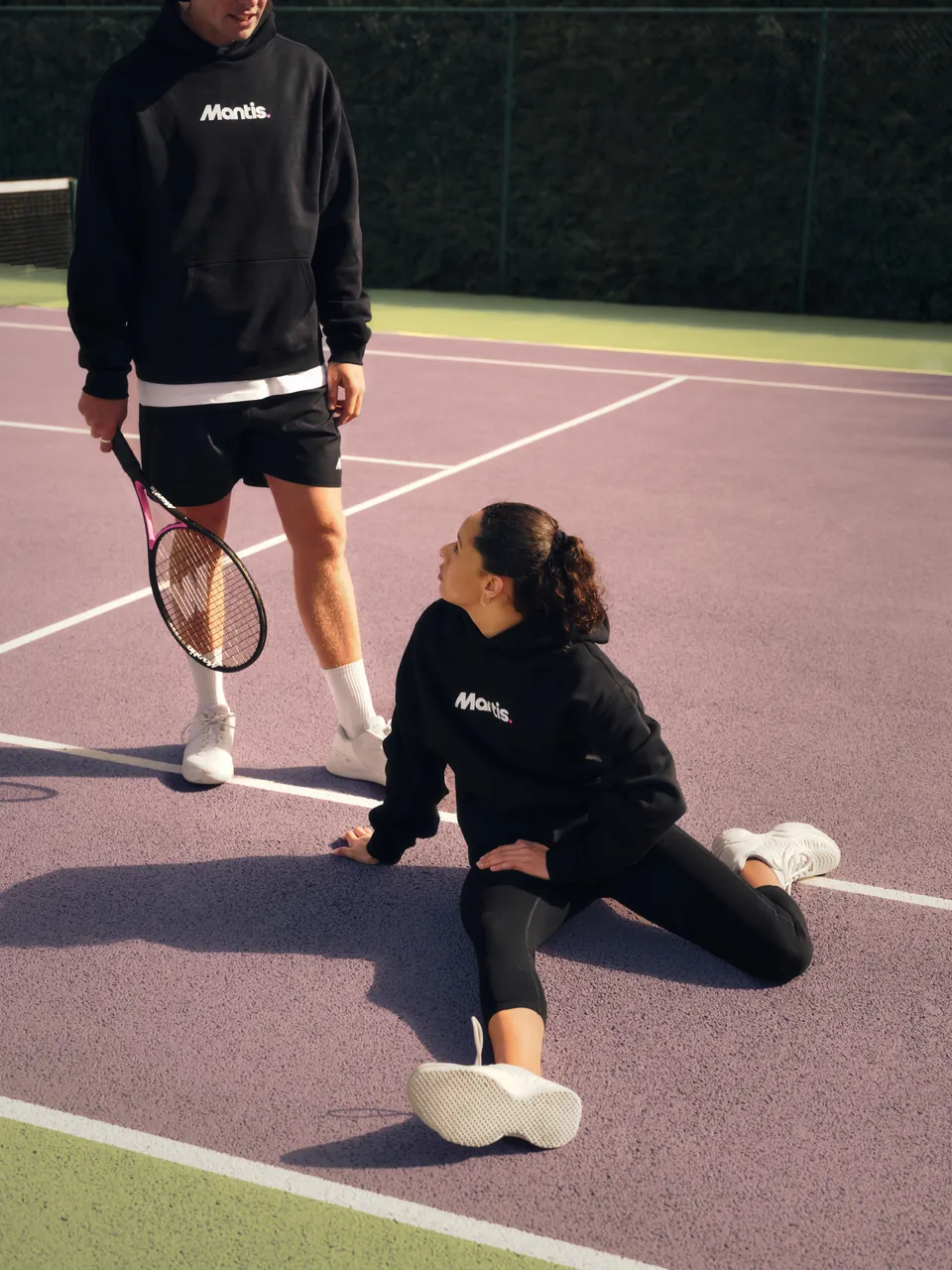 Two players on a tennis court in black Mantis kit, one stretching on the ground