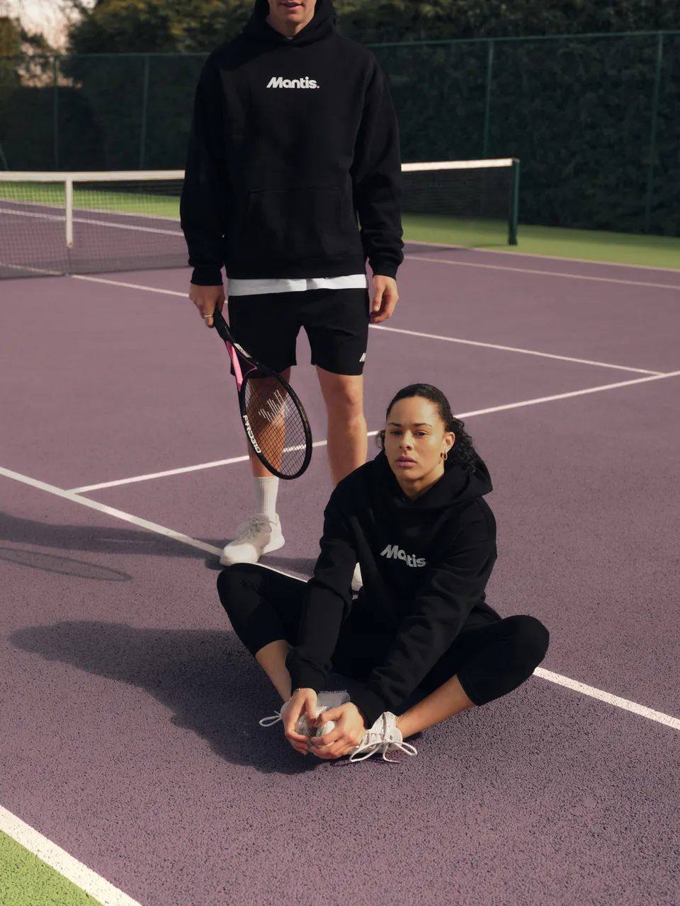 A player seated on the court in a black Mantis hoodie, another standing behind
